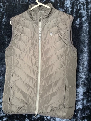 Grå dunväst från Emporio Armani EA7 - Snygg grå dunväst från Emporio Armani EA7 med dragkedja framtill och två fickor med dragkedja. Västen har ett diskret EA7-märke på bröstet och stilrena diagonala sömmar. Perfekt för lager-på-lager och streetwear. Äkthetslapp med Certilogo finns på insidan så att du enkelt kan verifiera plagget. 