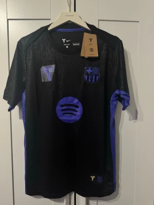 Svart FC Barcelona - Snygg svart FC Barcelona matchtröja från Nike med lila detaljer och klubbmärke på bröstet. tryckta loggor från Spotify och Kobe Bryant. Helt ny storlek M