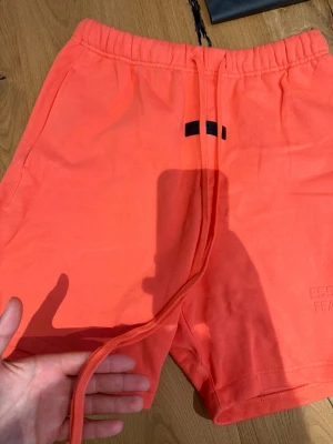 Orange shorts från Essentials Fear of God - Snygga orange shorts från Essentials Fear of God med elastisk midja och långa snören framtill. Tillverkade i mjuk bomull och har diskret logga på benet. Perfekta för en avslappnad och trendig look.