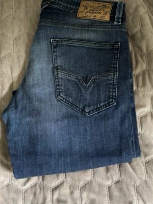 Diesel jeans med slitningar - Säljer ett par blå jeans med raka ben och snygga slitningar på låren. Jeansen har klassisk femficksdesign och en patch bak i midjan. Materialet är denim i bomull och färgen är mörkblå med ljusare tvättade partier.