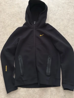 Svart Nike NOCTA hoodie med dragkedja - Svart hoodie från Nike NOCTA-kollektionen med gul logga på bröstet och detaljer på ärmen. Hoodien har hel dragkedja, två fickor med dragkedja och huva. Materialet är tjockt och mjukt, perfekt för en streetwear-look. Snygga gula broderade detaljer på ryggen.