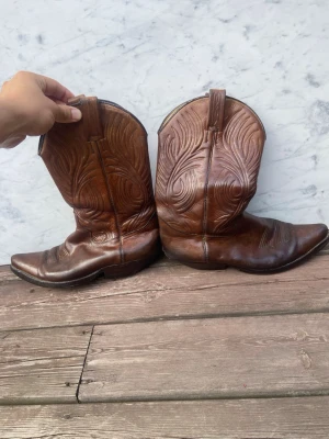 Bruna klassiska cowboy boots - Säljer ett par bruna cowboy boots i läder med snygga broderade detaljer längs skaftet. De har spetsig tå, låg klack och dragflikar upptill för enkel påtagning. Perfekta för dig som vill ha en tidlös westernstil. Skador synes på bilderna. Slitna på hälarna och lite vattenskada vid ett av hålen. 