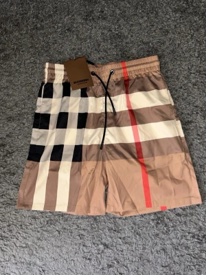 Rutig shorts från Burberry - Säljer ett par klassiska rutiga shorts från Burberry med beige, svart, vit och röd färg. Shortsen har snörning i midjan och den ikoniska Burberry-rutan. Perfekta för sommaren och riktigt snygga till en enkel t-shirt.