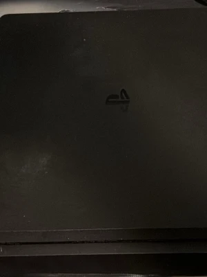PlayStation 4 spelkonsol svart - Säljer en svart PlayStation 4 spelkonsol från Sony. Konsolen har en stilren, rektangulär design med matt finish och PlayStation-loggan på ovansidan. Perfekt för gaming och streaming av filmer och serier.
