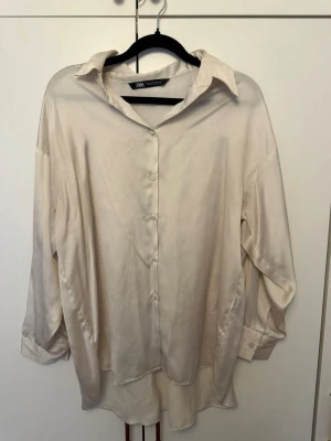 Krämvit skjorta från Zara - Stilren krämvit skjorta från Zara. Skjortan har klassisk krage, knäppning framtill och långa ärmar med knapp vid manschetten. Tillverkad i ett glansigt material som ger en lyxig känsla. Perfekt att styla till jeans eller kjol.