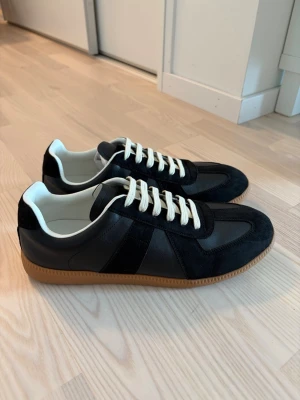 Svarta Maison Margiela sneakers - Snygga svarta sneakers med ovandel i både mocka och slätt läder, vita snören och klassisk gummisula i brunt. Skorna har en låg profil och stilren design som passar till det mesta. Insidan är vit och de har en tidlös look.