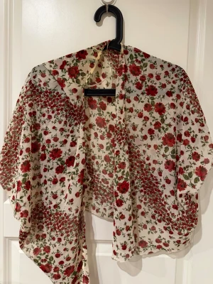 Blommig transparent bolerotopp H&M - Säljer en superfin bolerotopp i transparent syntetmaterial med blommigt mönster i rött och grönt på vit botten. Toppen har en loose och flowy passform, är ärmlös och perfekt för sommaren.
