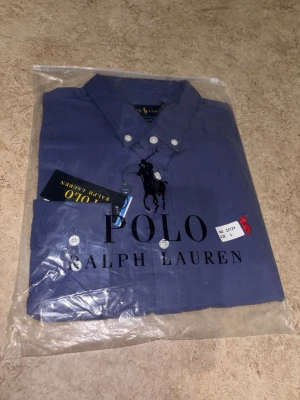 Blå skjorta från Polo Ralph Lauren - Klassisk blå skjorta från Polo Ralph Lauren med knappar framtill och button-down krage. Skjortan har en broderad logga på bröstet och är tillverkad i bomull för en bekväm känsla. Perfekt för en stilren och avslappnad look.