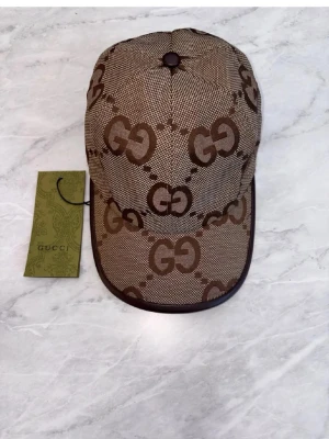 Beige keps med GG-mönster från Gucsi - Snygg beige keps från Gucsi med det klassiska GG-monogrammet i brunt över hela kepsen. Kepsen har böjd skärm, bruna detaljer och justerbar rem bak. Materialet är canvas med insida i bomull. Perfekt accessoar för dig som gillar exklusiv streetwear.