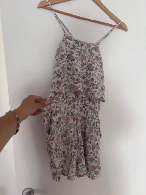 Blommig kort sommarklänning med short under - Söt kort klänning med tunna axelband och volangdetaljer. Klänningen har ett blommigt mönster i rosa, blått och vitt, perfekt för varma dagar. Tillverkad i ett lätt och luftigt material som känns skönt mot huden.