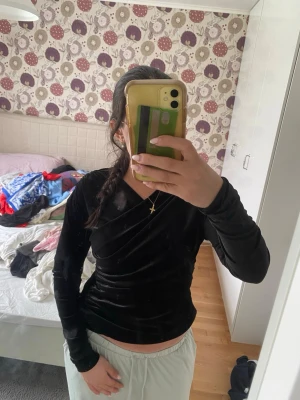 Svart omlottopp i sammet - Snygg svart omlottopp i mjuk sammet med lång ärm och draperad detalj på sidan. Toppen har en v-ringad omlottskärning som ger en stilren look och passar perfekt till jeans eller kjol. Märket är Gina Tricot.