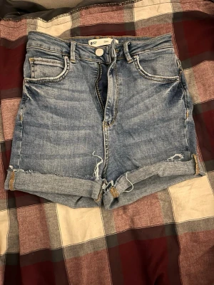 Blå denimshorts från Gina Tricot - Snygga blå denimshorts från Gina Tricot med hög midja och upprullade benslut. Klassisk femficksmodell med råa kanter och slitna detaljer för en avslappnad look. Perfekta för sommaren och enkla att matcha med allt.