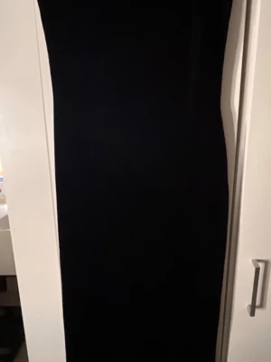 Svart långklänning med v-ringning - En stilren svart långklänning med v-ringning och ärmlös design. Klänningen har en rak och figurnära passform som ger en elegant siluett. Perfekt för dig som gillar minimalistisk och klassisk stil.