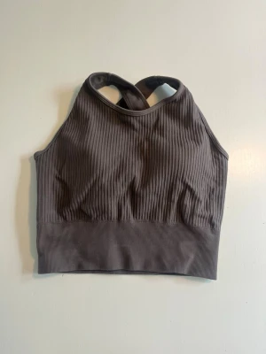 Ribbad brun linnetopp med korsrygg - Snygg ribbad brun linnetopp med korsade axelband i ryggen. Toppen har en sportig look och är tillverkad i stretchigt material som sitter tight mot kroppen. Perfekt för en trendig och avslappnad stil.