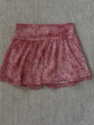 Rosa/röd mönstrad kortkjol  - Supersöt kortkjol i rosa, röd och vitt med paisleyinspirerat mönster. Kjolen har bred linning och ett lätt, luftigt tyg som ger en flowy känsla. Perfekt för varma dagar och enkel att styla med en enkel topp.
