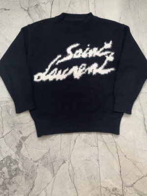 Svart stickad Saint Laurent-tröja - Cool svart stickad tröja från Saint Laurent med vit, suddig logotyp framtill. Tröjan har rund hals, långa ärmar och breda ribbstickade muddar vid ärmslut och nederkant. Perfekt för dig som gillar streetstyle och exklusiva märken.