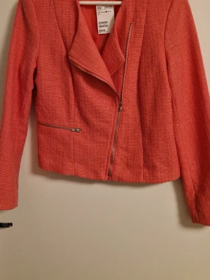 Korallrosa kavaj med dragkedja H&M - Snygg korallrosa kavaj från H&M med asymmetrisk dragkedja och en ficka med dragkedja. Kavajen har en strukturerad, vävd yta och är figursydd med långa ärmar. Perfekt för att lyfta din outfit med färg och stil.