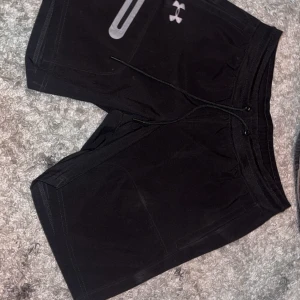 Svarta träningsshorts från Under Armour - Svarta träningsshorts från Under Armour med snörning i midjan och en praktisk ficka med dragkedja framtill. Perfekta för gymmet eller löprundan, med bekväm passform och sportig look.
