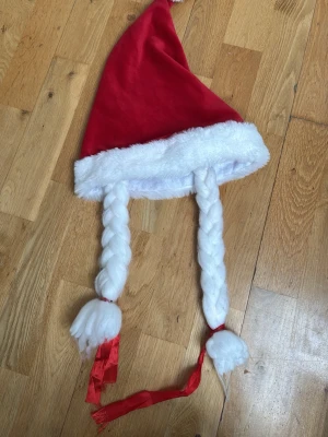 Tomteluva med flätor och rosetter - Rolig tomteluva i röd sammet med vit fluffig kant och två långa vita flätor med röda rosetter. Perfekt för maskerad eller juliga cosplay outfits. Flätorna är fastsydda och luvan har klassisk spetsig form.