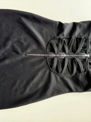 Svart kort klänning med snörning - Snygg svart kort klänning med halterneck och cool snörningsdetalj framtill. Klänningen har dragkedja och är tillverkad i ett stretchigt material som sitter tight. Perfekt för dig som vill ha en trendig och edgy look.