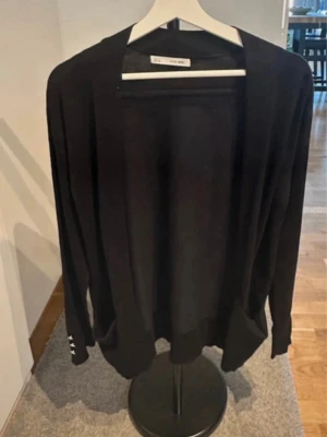 Svart öppen kofta  - Stilren svart öppen kofta med lång ärm och enkel design. Koftan är tunn och mjuk, perfekt att slänga över en t-shirt eller topp. Klassisk modell utan knappar och med diskreta detaljer vid ärmslutet.