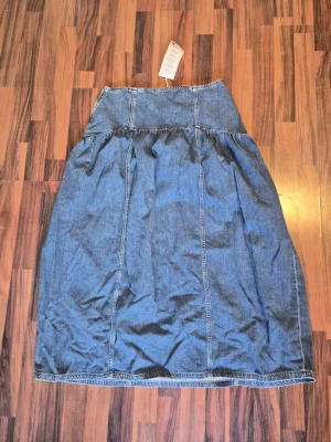 Blå lång jeanskjol från NA-KD - oanvänd! - Säljer en helt ny blå lång jeanskjol från NA-KD med hög midja och klassisk denimlook. Kjolen har synliga sömmar och en lätt utställd passform som ger en avslappnad vibe. Perfekt att styla med sneakers eller boots för en trendig look. Helt ny oanvänd!