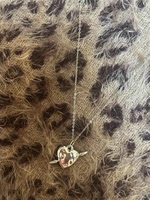 Silverhalsband med hjärthänge - Snyggt halsband i silverfärg med ett hjärthänge som har en pil genom sig. Kedjan är tunn och smidig, perfekt för att ge en cool touch till din outfit. Hjärtat har en blank yta som reflekterar ljuset snyggt. Rostfri 
