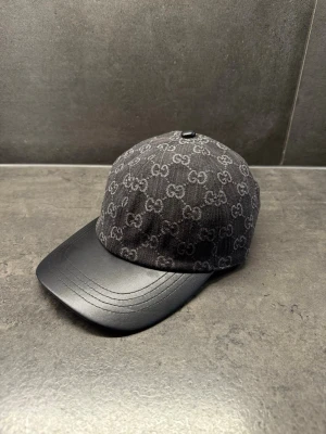 Svart keps med GG-mönster - Snygg svart keps från med klassiskt GG-monogram över hela tyget och böjd skärm i svart läder. Kepsen har en modern och stilren look som passar perfekt till streetwear och modeintresserade. Materialmix av textil och läder ger en exklusiv känsla.    LÄGSTA PRIS 520