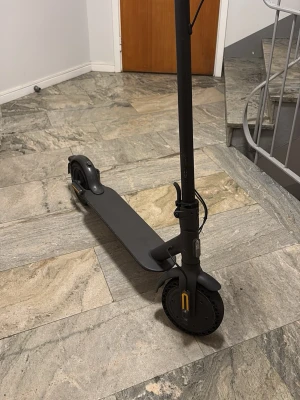 Svart elscooter med gråa handtag - Elscooter i svart med gråa handtag och robust ram. Utrustad med stora hjul, handbroms och reflexdetaljer framtill. Scootern har en minimalistisk design och är tillverkad i metall och plast. Perfekt för smidig transport i stan.