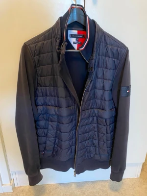 Mörkblå quiltad jacka Tommy Hilfiger - Snygg mörkblå jacka från Tommy Hilfiger med quiltad front och ärmar i mjuk bomullsmix. Jackan har dragkedja, ståkrage med rödvit detalj och logga på ärmen. Perfekt för dig som gillar stilren och sportig look.