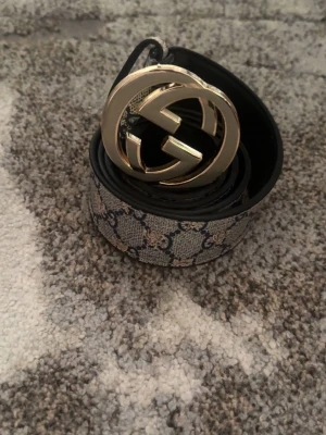 Gucci GG - Snyggt Gucci GG Supreme bälte med klassiskt guldigt GG-spänne. Bältet har ett grått och svart mönster med GG-loggor och är gjort i skinn och canvas. Perfekt accessoar för att lyfta din outfit med en lyxig touch. Pris kan diskuteras. Köpt för 900kr