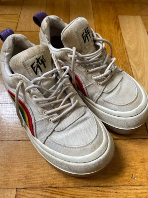Eytys vita sneakers - Snygga vita sneakers från Eytys med regnbågsfärgad detalj på sidan. Skorna har en chunky sula, lila hälflik och är tillverkade i textil med gummisula. De är använda och hälen är sliten därav lägre pris!