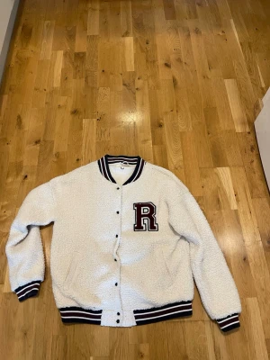 Cubus Vit teddyjacka med R-märke stl M  - Snygg vit teddyjacka i collegestil med svart och vinröda detaljer på krage, ärmslut och midja. Stor R-patch i vinrött på bröstet. Jackan har knäppning framtill och två fickor. Perfekt för dig som gillar sportig och trendig stil.