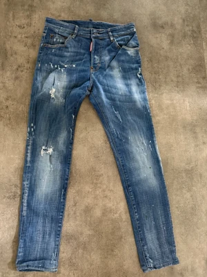 Blå jeans från Dsquared2 - Säljer ett par blå jeans från Dsquared2 med coola slitningar och distressed detaljer. Jeansen har smal passform, klassisk femficksdesign och knappgylf. Materialet är denim i bomull och de har en casual vibe med unika slitningar och färgstänk.