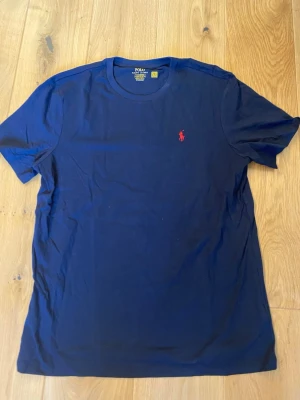 Svart t-shirt från Polo Ralph Lauren - Snygg Svart t-shirt från Polo Ralph Lauren med klassisk röd logga broderad på bröstet. T-shirten har rund halsringning och korta ärmar. Tillverkad i mjuk bomull som känns skön mot huden. Perfekt basplagg för en clean och stilren look.