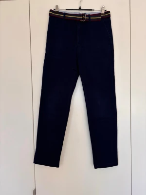 Mörkblå chinos med randigt bälte, Polo Ralph Lauren, strlk 14 år - Snygga mörkblå chinos med raka ben och ett färgglatt randigt bälte i midjan. Klassisk passform med fickor fram och bak. Perfekta för dig som vill ha en stilren men ändå lekfull look. Materialet ser ut att vara bomull eller bomullsblandning.