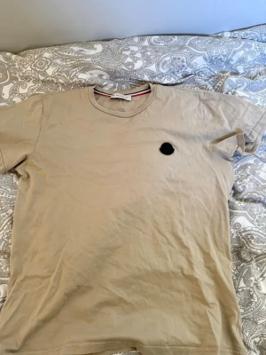 Beige Moncler t-shirt med logga - Snygg beige t-shirt från Moncler med klassisk rund halsringning och liten svart logga på bröstet. T-shirten har korta ärmar och är tillverkad i mjuk bomull. Perfekt för en clean och stilren look.
