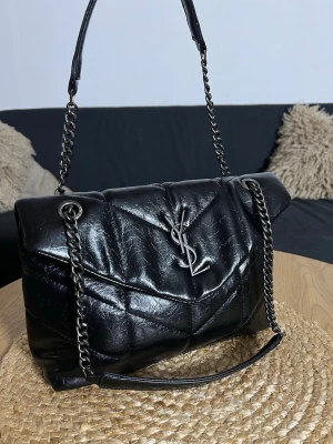 Svart YSL handväska  - Snygg svart handväska från Saint Laurent i quiltat läder med silverfärgad YSL-logga framtill. Väskan har kedjeremmar i metall och en mjuk, puffig form. Perfekt för dig som vill ha en lyxig och trendig accessoar till din outfit.