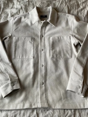 Overshirt från Jack&Jones - Stilren ljusgrå overshirt från Madewell med två stora bröstfickor och tryckknappar framtill. Overshirten har klassisk krage och raka ärmar med knapp vid ärmslut. Tillverkad i ett mjukt, tjockare tyg som ger en clean look. Passar även M och är bara använd ett fåtal gånger.