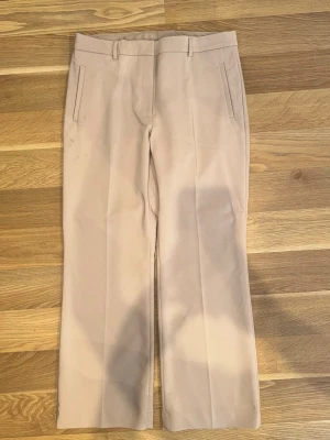 Beige kostymbyxa med raka ben  - Snygga beige kostymbyxor med raka ben och pressveck. Byxorna har hällor för skärp, två sidofickor fram och två passpoalfickor bak. Perfekta för en stilren och clean look. Materialet känns mjukt och har en lätt stretch.