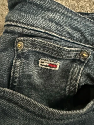 Blå jeans från Tommy Hilfiger - Säljer ett par klassiska blå jeans från Tommy Hilfiger med snygga detaljer och broderad logga på fickan. Jeansen har fem fickor, slitstarka nitar och en mörk tvätt med lätt slitning. Perfekt för dig som gillar stilrena och tidlösa plagg.