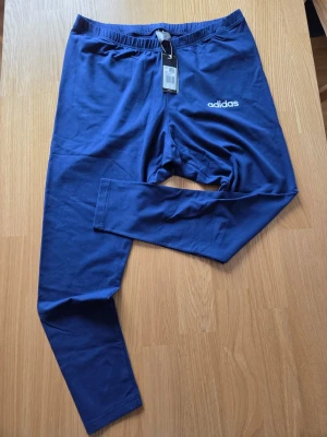 Adidas Essentials Linear tights , leggings , storlek 3XL - Artikelnummer : IS6970  Ett par mörkblå tights från Adidas med vit logotyp på vänster ben. Tightsen har en resår i midjan och en åtsittande passform.  Ett enkelt och sofistikerat utseende. Dessa leggings adidas är ett måste för mysiga kvällar eller hektiska dagar. De är tillverkade av bomull med en antydan till stretch och gör att du kan röra dig fritt vid alla tillfällen. Den höga midjan smickrar din figur och ger optimalt stöd. De passar perfekt med en beskuren tröja eller ett överdimensionera