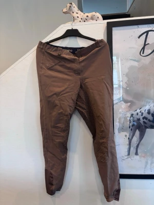 Bruna ridbyxor från Domino - Snygga bruna ridbyxor från Domino med förstärkning på insidan av benen. Byxorna är tillverkade i en mix av polyester, bomull och elastan för extra komfort och stretch. Klassisk design med fickdetalj framtill och smal passform.