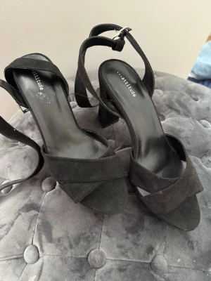 Svarta klackar från Attitude - Svarta pumps från Attitude med bred klack och öppen tå. Skorna har en korsad rem över foten och justerbart spänne vid ankeln. Materialet är mockaimitation. 