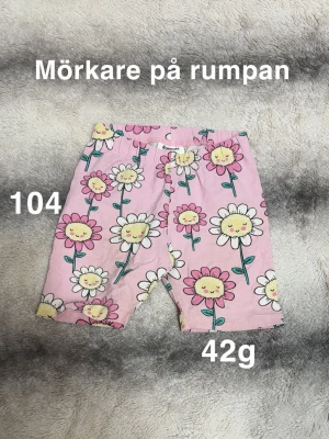 Rosa blommiga shorts från Kappahl - Säljer ett par söta rosa shorts från Kappahl med färgglada blommor och glada ansikten. Shortsen har resår i midjan. Perfekta för en lekfull och färgstark stil i sommar🌸 Mörkare på rumpan. Storlek 104