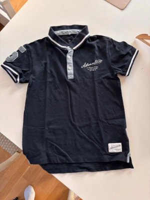Mörkblå piké t-shirt med tryck, 146/152 - Snygg mörkblå piké t-shirt från name it med vit text på bröstet och patch på ärmen. T-shirten har korta ärmar, vit krage och knappar framtill. Materialet är mjuk bomull och detaljer i vitt vid ärmslut och krage ger en sportig look.