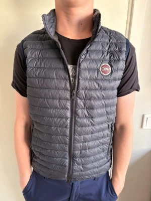 Mörkblå väst från Colmar - Snygg mörkgrå dunväst från Colmar med quiltad design och hög krage. Västen har dragkedja framtill, två sidofickor med dragkedja och en rund Colmar-logga på bröstet. Perfekt för lager-på-lager och streetstyle. Mycket bra skick! Storlek 160-170 cm. Modellen är 172 cm