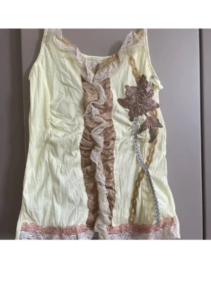 Gul spetslinne med volang och brodyr - Snyggt ljusgult linne med tunna axelband, volang i beige och spetsdetaljer framtill. Dekorerad med broderad blomma och spets längs nederkanten. Perfekt för en unik och feminin stil.