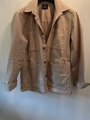Beige overshirt från Less Worries - Stilren beige overshirt från Less Worries med klassisk krage och knäppning framtill. Fyra stora fickor framtill ger en cool utility-look. Tillverkad i ett mjukt bomullstyg som känns skönt mot huden. Perfekt lager-på-lager-plagg för en avslappnad stil.