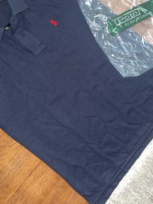 Marinblå polo t-shirt från Polo Ralph Lauren - Marinblå polo t-shirt från Polo Ralph Lauren med klassisk krage, knappar vid halsen och röd broderad logga på bröstet. Tillverkad i mjuk bomull och har korta ärmar för en clean och tidlös stil.
